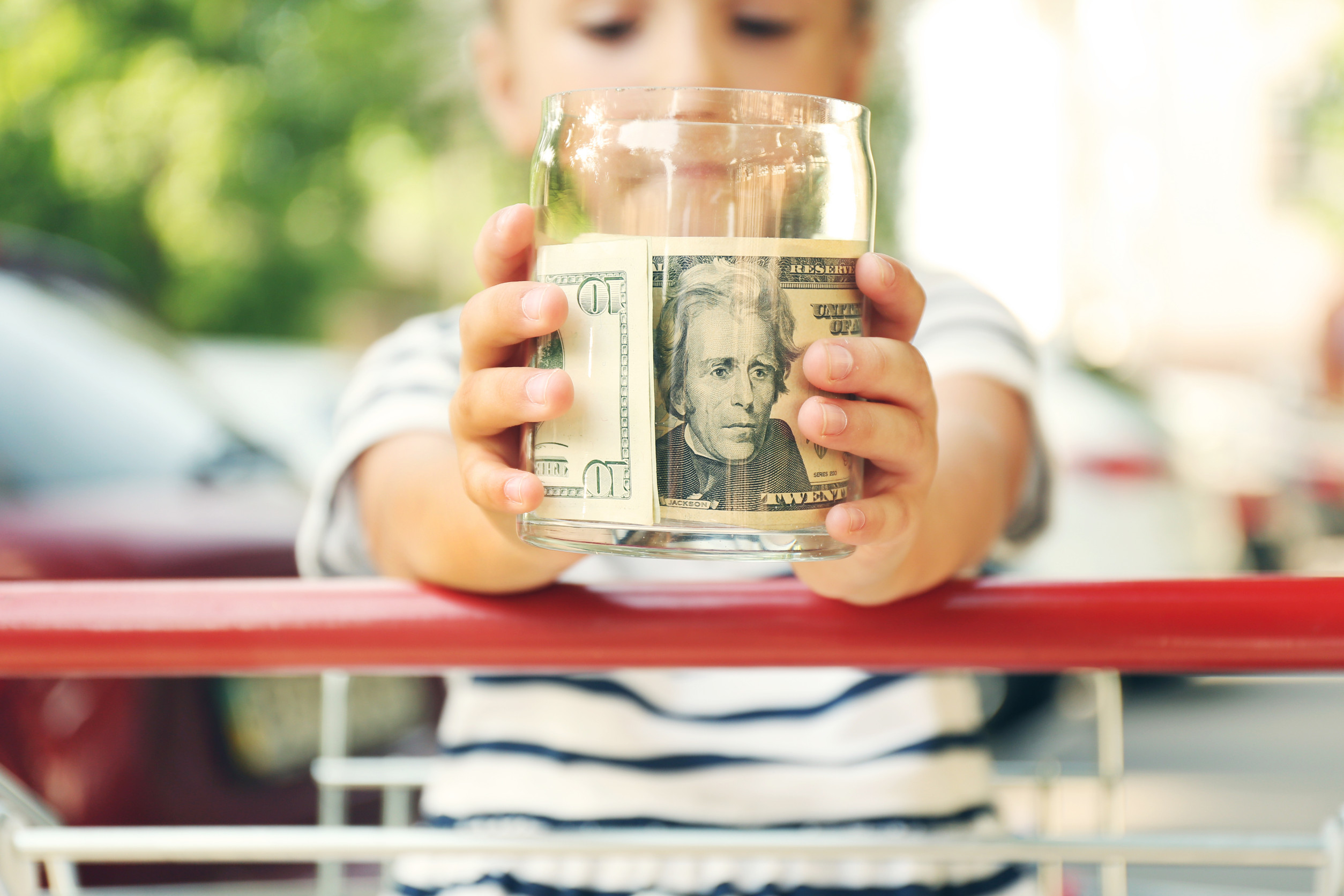 Cute girl holding bank with money in hands and sitting in shopping cart, outdoors