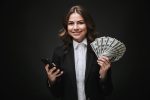 Portrait of a confident young businesswoman wearing formal suit standing isolated over black background, holding mobile phone, showing money banknotes