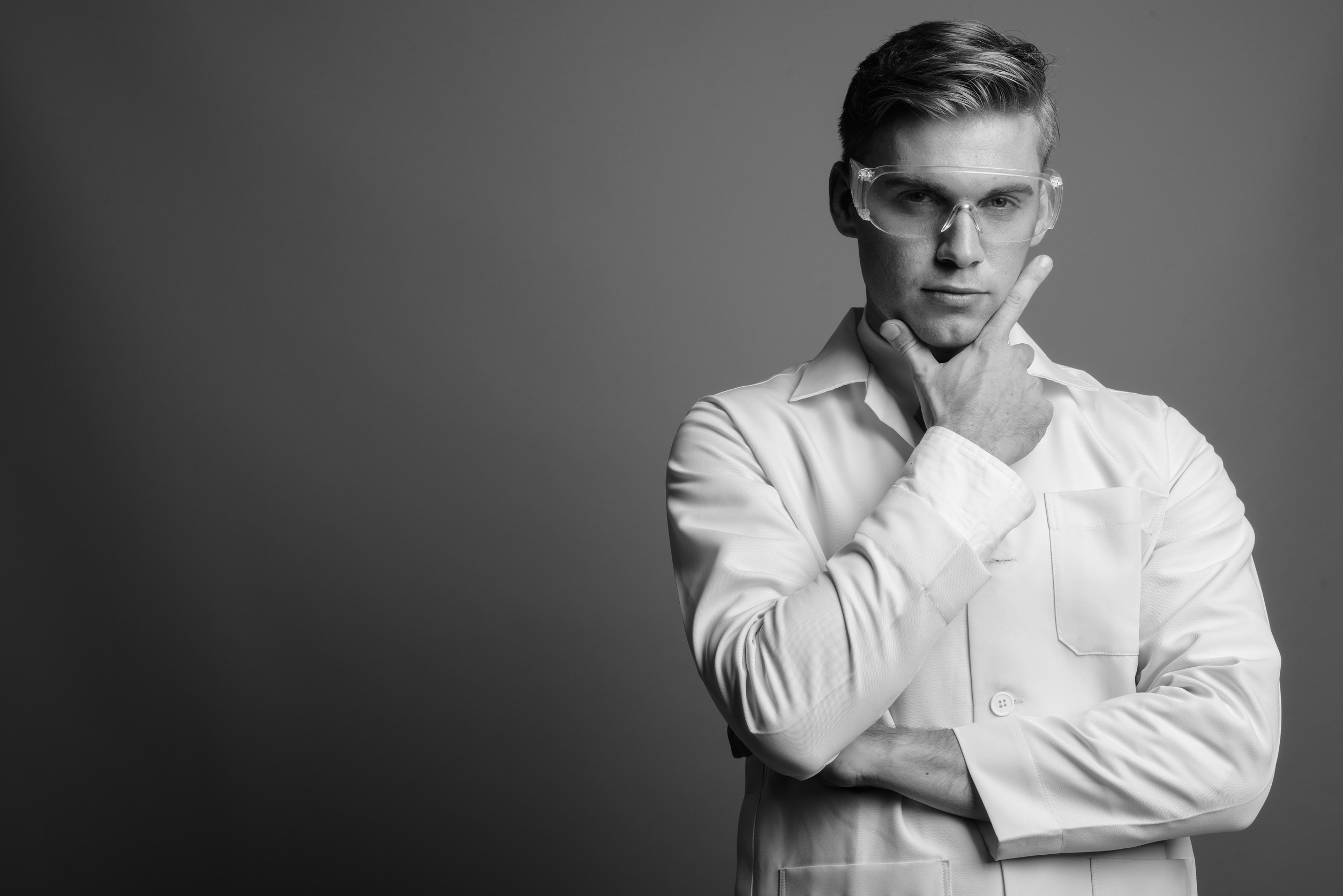 Studio shot of young handsome man doctor as scientist wearing protective glasses against gray background in black and white