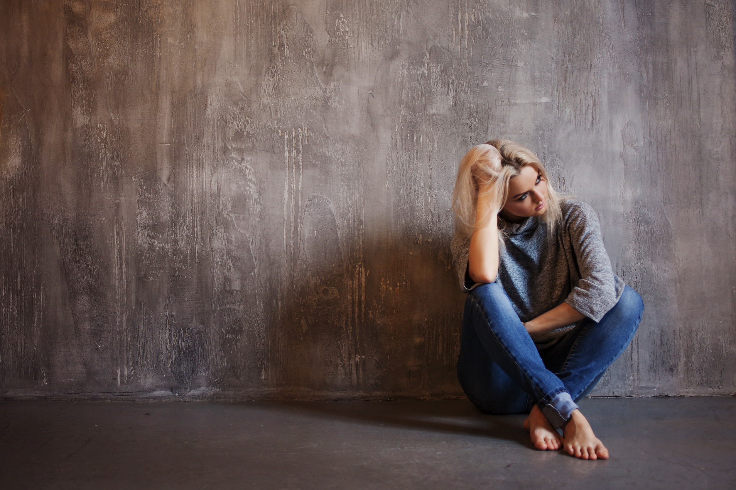 Sad woman sits on the floor. Depression and chronic fatigue. Young beautiful blonde in a gray sweater and jeans, gray textured background