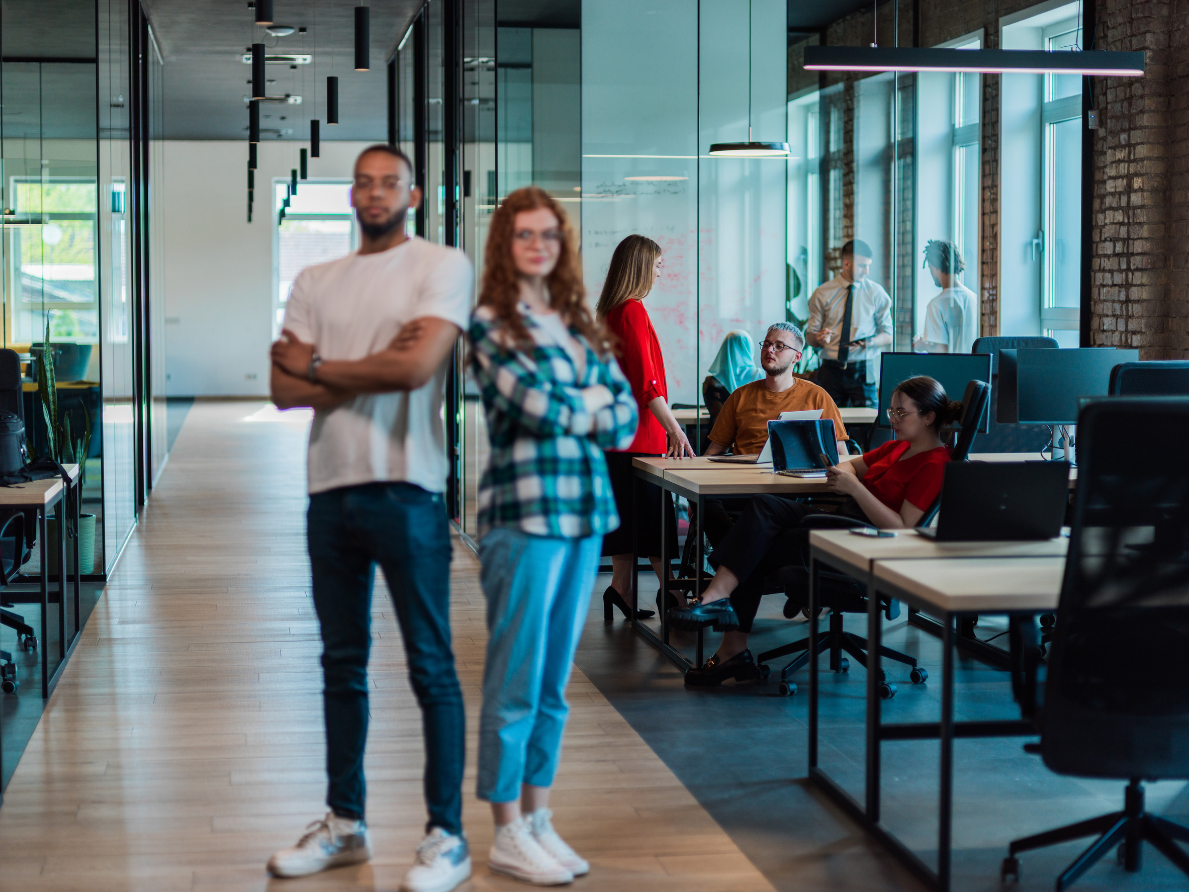 African-American businessman and his businesswoman colleague stand at the forefront with crossed arms, exuding confidence and leadership, while their diverse team diligently works behind them, symbolizing teamwork and corporate success in a modern and inclusive workplace.