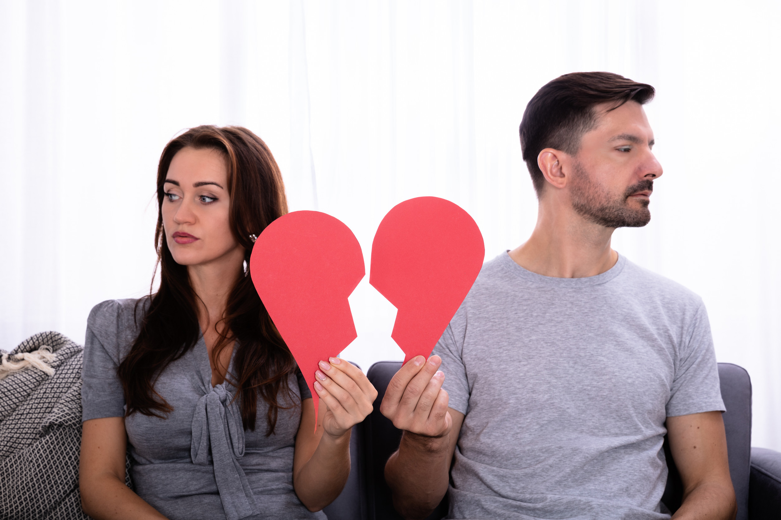 Couple Sitting On Sofa Ignoring Each Other And Holding Broken Red Heart