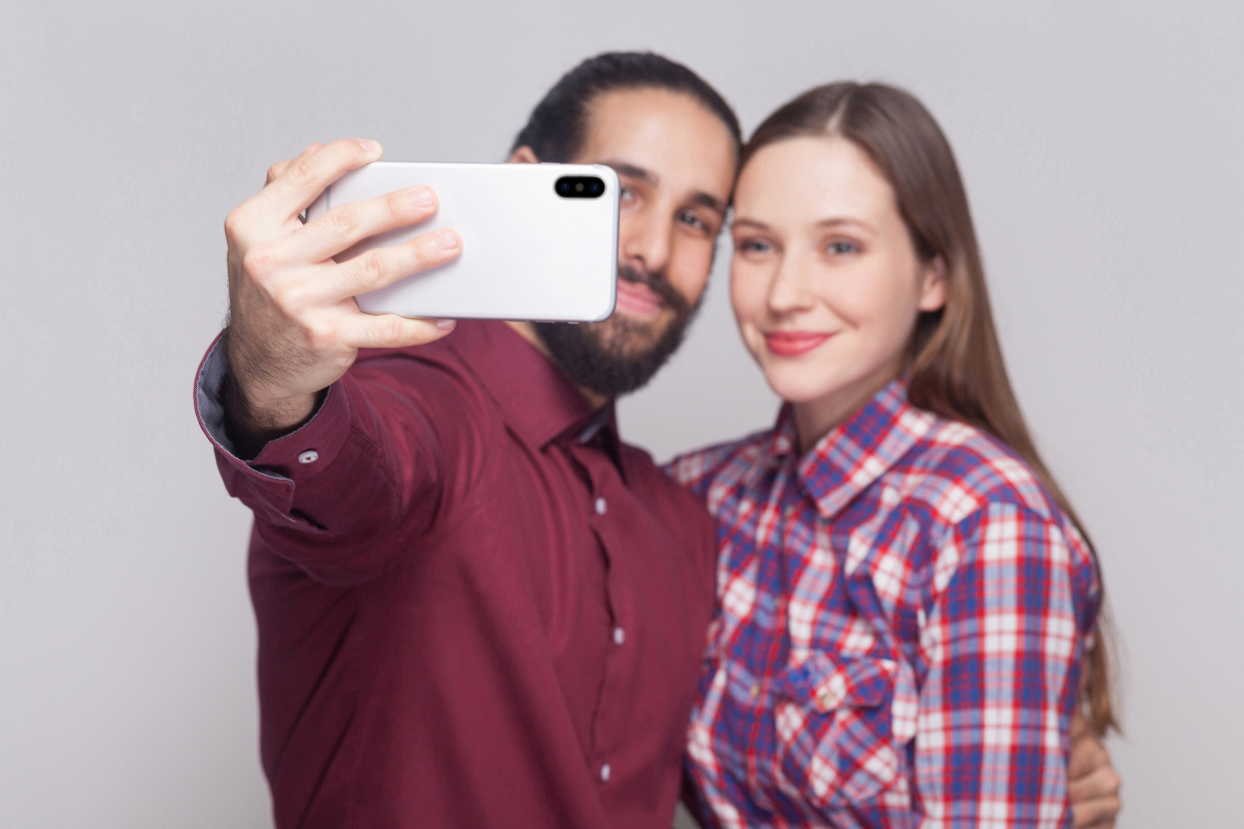 Portrait of happy satisfied couple standing, looking and smiling at smartphone camera to make selfie or video call. focus on mobile smart phone. indoor studio shot, isolated on grey background.